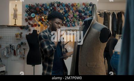 African American fashion designer stands near the mannequin in unfinished tailored wedding or business suit with digital tablet computer and thinks. Seamstresses working on background in atelier. Stock Photo