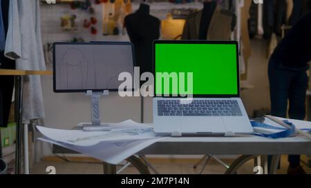Dolly shot of laptop with green screen and digital tablet computer with sketch on table in atelier workshop. Tailor and seamstress work on background. Concept of fashion and technologies in business. Stock Photo
