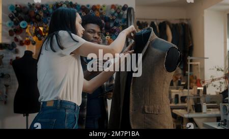 African American fashion designer talks with seamstress in atelier and shows sketch of future suit on digital tablet computer. Mannequin in unfinished tailored suit. Concept of fashion and hand craft. Stock Photo