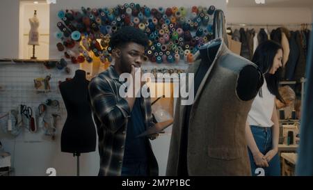 African American fashion designer stands near the mannequin in unfinished tailored wedding or business suit with digital tablet computer and thinks. Seamstresses working on background in atelier. Stock Photo