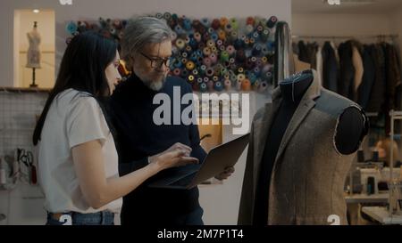 Male mature fashion designer shows sketch of future jacket to seamstress on laptop in atelier workshop and discusses tailoring process. Mannequin in unfinished suit. Fashion and hand craft concept. Stock Photo