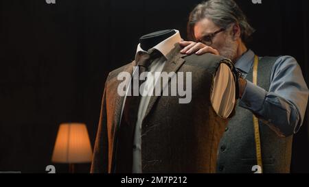 Male mature tailor works on elegant business or wedding suit. Mannequin with tailored shirt, tie and jacket in stylish luxury designer atelier or tailoring dim studio. Fashion and hand craft concept. Stock Photo