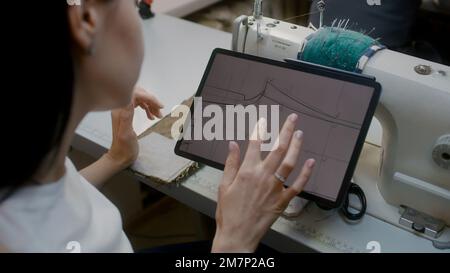Female tailor sits at sewing machine with digital tablet and zooming picture. Dressmaker or seamstress watches sizes of custom apparel at sketch and sewing suit. Atelier workshop. Concept of fashion. Stock Photo