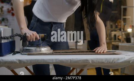 Woman irons fabric textile in atelier workshop. Her colleagues working on background: man works with tailored suit, seamstress works at sewing machine. Concept of fashion and hand craft. Stock Photo