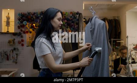 Seamstress irons new tailored shirt with electric hand steam generator in atelier workshop. Male tailor sews clothes on background. Concept of small business, tailoring industry and fashion. Stock Photo