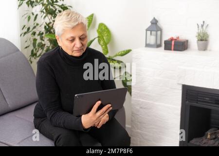 Smiling middle-aged Caucasian woman sit on couch in living room browsing wireless Internet on tablet, happy modern senior female relax on sofa at home Stock Photo