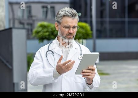 Serious mature doctor in medical gown outside modern clinic using tablet computer for video call, man with stethoscope consults patient online remotely. Stock Photo