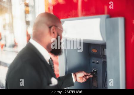 A shallow depth-of-field shot with a selective focus on an elegant hairless black man's hand, inserting a credit card into the red and white ATM machi Stock Photo