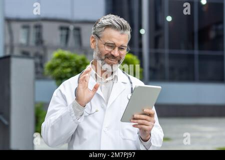 Senior gray-haired smiling doctor talking to patients remotely, a man in a medical coat and a stethoscope holds a tablet computer in his hands, consults patients online from outside a modern clinic. Stock Photo