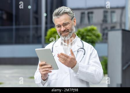 Senior gray-haired smiling doctor talking to patients remotely, a man in a medical coat and a stethoscope holds a tablet computer in his hands, consults patients online from outside a modern clinic. Stock Photo