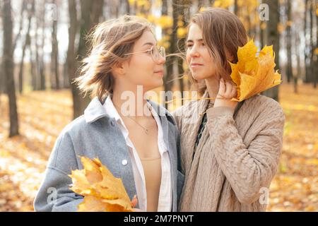 Girls look to each other and hold bouquet of fallen yellow maple leaves. Happy mother and daughter walk in autumn park.
