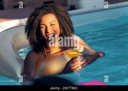 A young multiethnic woman with curly hair is laughing and having a great time with her friends, as she holds a martini cocktail floating on a donut-sh Stock Photo