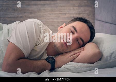 sleeping beautiful man. A satisfied expression on his face. A young guy is resting in bed with a smartwatch on his hands. Stock Photo