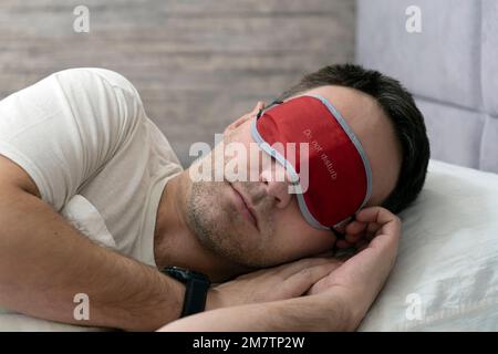 Calm caucasian young man in sleep mask white t-shirt lying in bed sleeping slumber resting with closed eyes relaxing at home indoors bedroom. Good moo Stock Photo