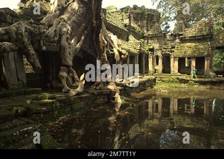 A woman visitor walking on a walkway between an ancient man-made pond and the ruins of Preah Khan temple in Siem Reap, Cambodia. Ponds, canals and reservoirs in Angkor Archaeological Park were constructed to support an ancient civilization of Khmer empire, which was the biggest and most populated city in the world where up to 900,000 people lived, until wars and climate catastrophe in 14th and 15 centuries had forced it to be abandoned. Stock Photo