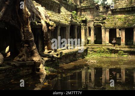 A woman walking on a walkway between an ancient man-made pond and the ruins of Preah Khan temple in Siem Reap, Cambodia. Ponds, canals and reservoirs in Angkor Archaeological Park were constructed to support an ancient civilization of Khmer empire, which was the biggest and most populated city in the world where up to 900,000 people lived, until wars and climate catastrophe in 14th and 15 centuries had forced it to be abandoned. Stock Photo