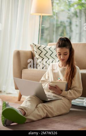 Young woman watching educational video on laptop and taking notes in planner Stock Photo