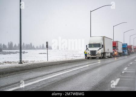 Professional truck driver is installing chains on semi truck standing on the road shoulder side in a convoy of big rigs semi trucks with semi trailers Stock Photo