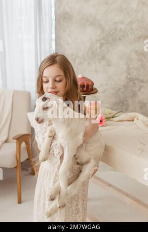 The girl is sitting at the Easter table with cakes, spring flowers and quail eggs and holding a white kid in her arms. Happy Easter celebration Stock Photo