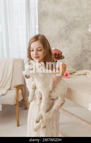 The girl is sitting at the Easter table with cakes, spring flowers and quail eggs and holding a white kid in her arms. Happy Easter celebration Stock Photo