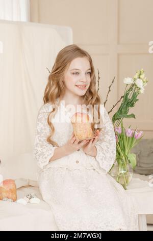 A girl with long hair in a light dress is sitting at the Easter table with cakes, spring flowers and quail eggs. Happy Easter celebration Stock Photo