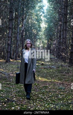 A young blonde women in long green coat walking down a path between trees in a forest, holding a gun and a briefcase. Thriller book cover style. Stock Photo