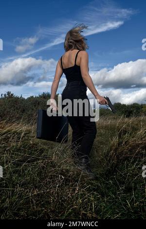 A young blonde woman wearing black holding a gun (pistol) and a briefcase in grassland near woods. Thriller book cover style. Stock Photo