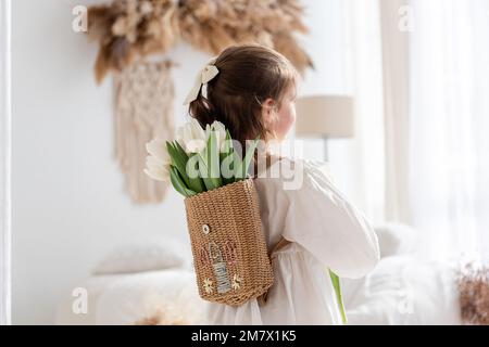 Little girl in white dress, nightgown stands with back, faceless, on shoulders is hand-made backpack made of raffia with tulips. Light airy, spring in Stock Photo