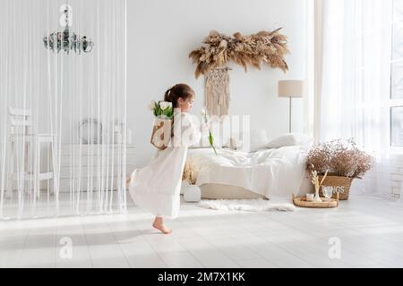 Little girl in white dress, nightgown dancing with back, faceless, on shoulders is hand-made backpack made of raffia with tulips. Light airy, spring i Stock Photo