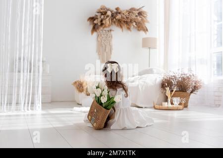 Little girl in white dress, nightgown sitting with back, faceless, on shoulders is hand-made backpack made of raffia with tulips. Light airy, spring i Stock Photo