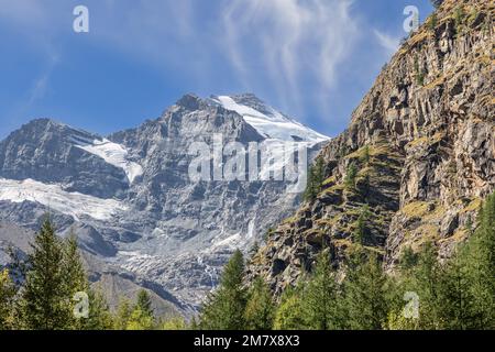Gorge overgrown with green coniferous forest and steep granite rocks rising from it covered with snow on peaks in Gran Paradiso National Park, Italy Stock Photo