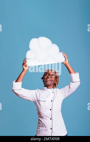 Cheerful woman wearing chef jacket holding cloud of text above head with space for advertising. Smiling cook with white text bubble in studio shot against blue background. Stock Photo