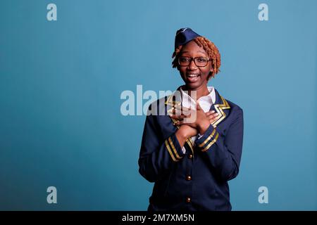 Happily smiling african american stewardess lady portrait. Female flight worker looking surprised, expectant, glad at camera. Suit dressed lucky woman laughing on blue background. Stock Photo