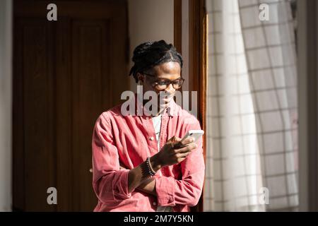 Happy glad African American hipster man in eyeglasses using smartphone standing near window at home. Stock Photo