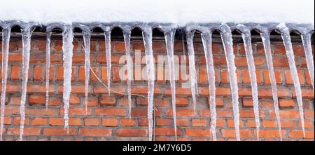 Snow-covered roof of a house with icicles close-up on background of red brick wall Stock Photo