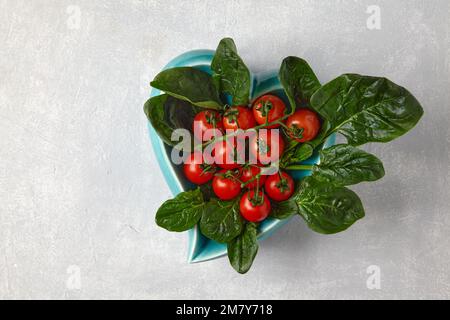 Red cherry tomatoes on a branch and green spinach leaves in a heart-shaped blue bowl on a light concrete table Stock Photo