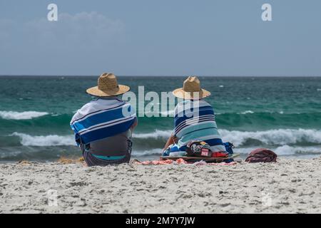 A couple sat on a beach wrapped in striped towels and wearing matching sun hats looking out to sea Stock Photo