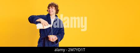Smiling young man in bathrobe pouring milk into bowl isolated over bright yellow background. Positive emotions, home cozy atmosphere, funny meme Stock Photo