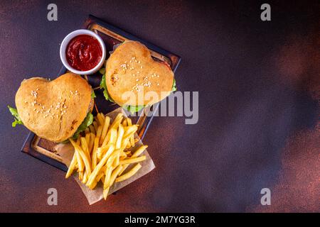 Valentine day Heart-shaped burgers. Two tasty cheeseburgers with french fries and beer bottles on dark table background. Idea for Valentine day dinner Stock Photo