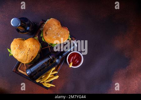 Valentine day Heart-shaped burgers. Two tasty cheeseburgers with french fries and beer bottles on dark table background. Idea for Valentine day dinner Stock Photo