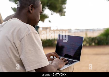 Black man typing on laptop keyboard and creating code while working on freelance project on beach in summer Stock Photo