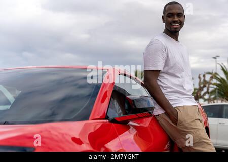 Cheerful African American male driver in casual clothes smiling and looking at camera while leaning on red car Stock Photo