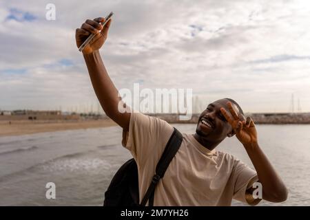 Cheerful young African American male in casual clothes taking selfie on smartphone while showing victory sign near sea at beach Stock Photo