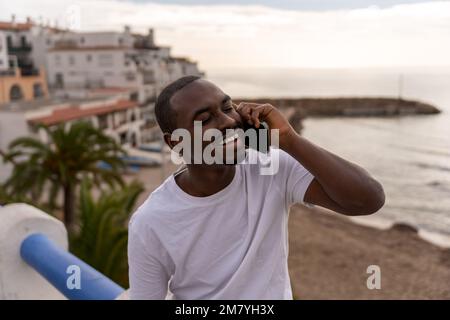 Positive African American male in casual clothes smiling and talking on smartphone while relaxing near sea during vacation Stock Photo