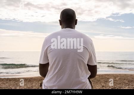 Low angle back view of unrecognizable African American male in casual white t shirt sitting on sandy beach and admiring waving sea Stock Photo