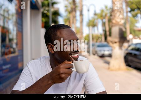 Smiling young bearded African American man in casual clothes sitting at table in outdoor cafe and drinking hot coffee while enjoying cityscape Stock Photo