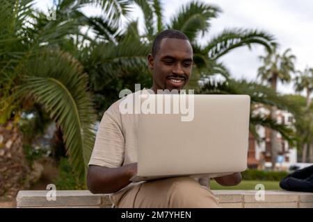 African American male programmer in casual clothes smiling and browsing laptop while sitting on border near palm trees and working remotely  Stock Photo