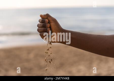 Crop anonymous African American male pouring sand from hand during day on beach near waving sea Stock Photo