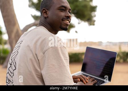 Positive African American male programmer in t shirt smiling and looking away while using netbook during remote work on beach on summer day Stock Photo