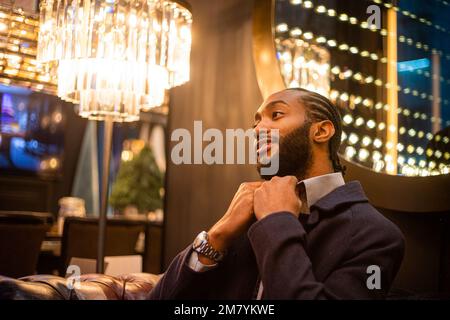 An attractive African man with pigtails straightens his shirt while sitting on a sofa Stock Photo